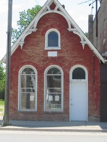 Front view of the Wardsville Museum. The structure is red brick, with two arched windows on the left and an arched door on the right. In the centre of the gable there is a smaller arched window above the middle lower window. The gable has white gingerbread trim on the upper half